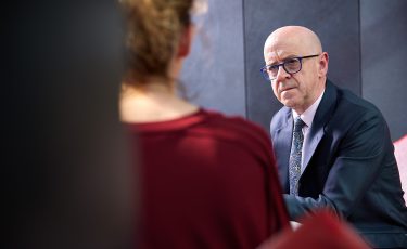 A man in blue glasses and a navy suit looks towards the camera. He's in discussion with a woman in a red dress.