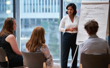 Core Skills in Coaching & Mentoring for Leaders and Managers. Woman teaches two others, writing on a paper whiteboard.